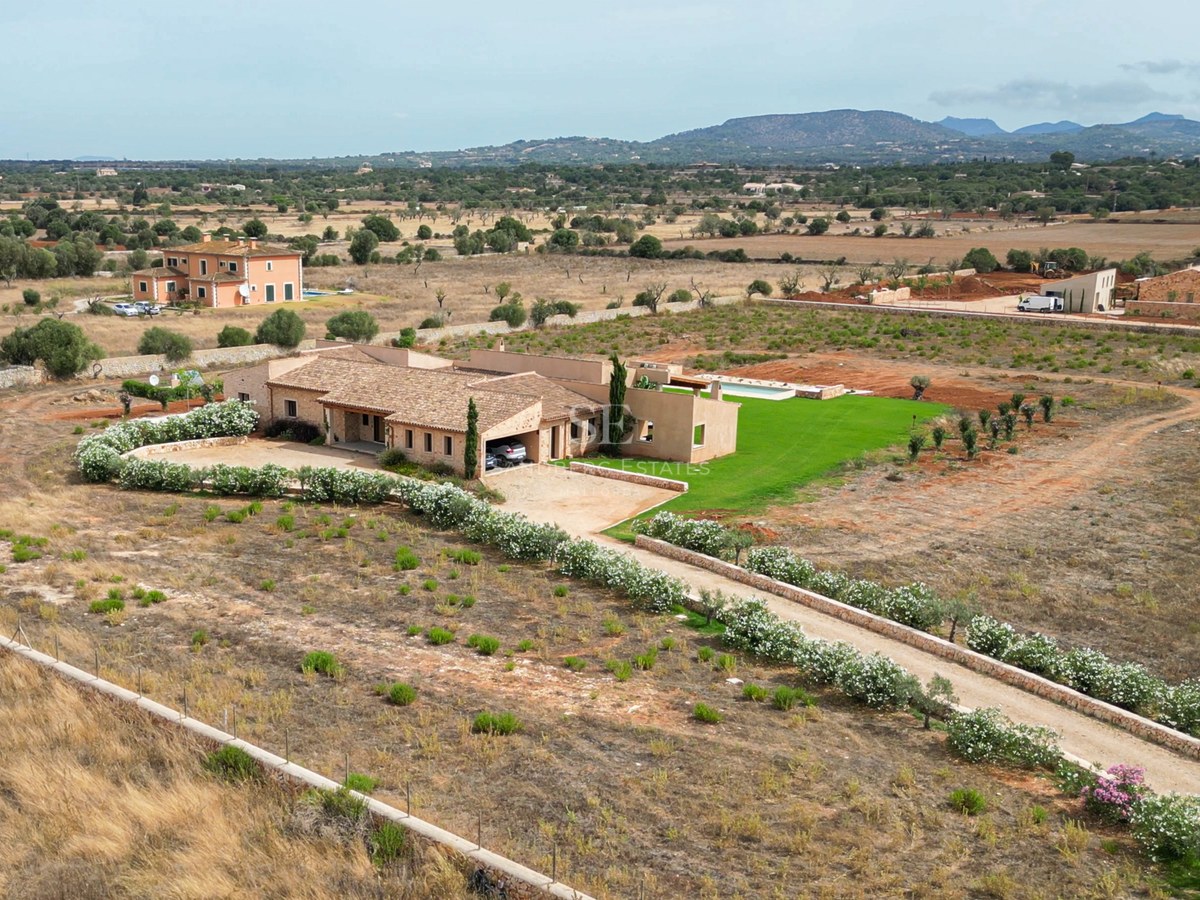 Aerial shot of a stone villa with a pool and flower-lined driveway in a Mediterranean countryside setting.