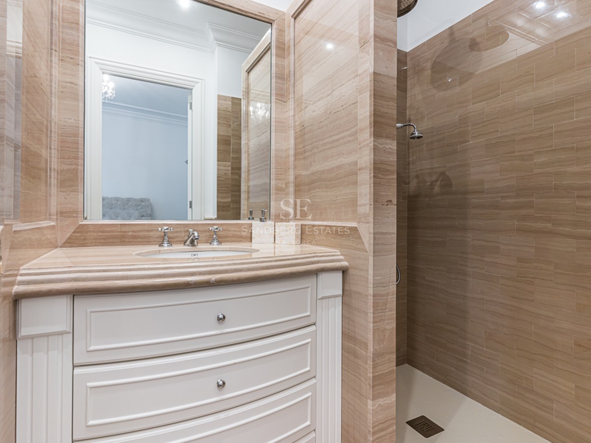 Luxury bathroom featuring beige travertine tile walls, a white curved vanity with marble top, and a walk-in shower.