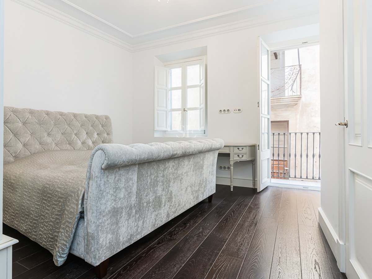 Bright bedroom featuring a grey velvet tufted bed, dark wood floors, and a balcony with wrought iron railing.