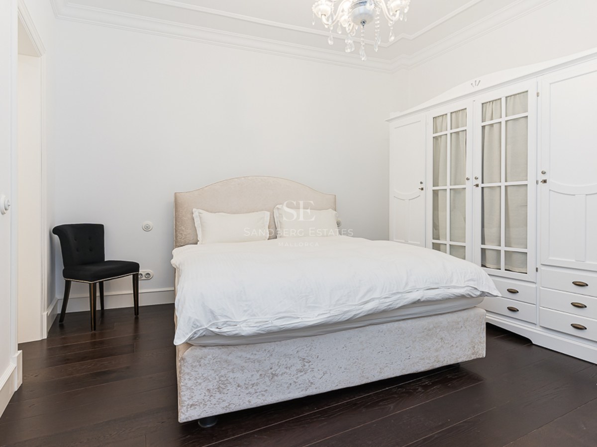 Bright bedroom featuring a beige bed, large white wardrobe, black chair, and crystal chandelier on dark wood floors.