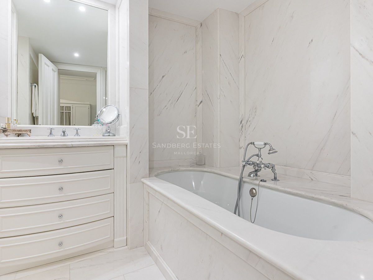 Bright master bathroom featuring white marble walls, a built-in tub with chrome faucets, and a white vanity with drawers.
