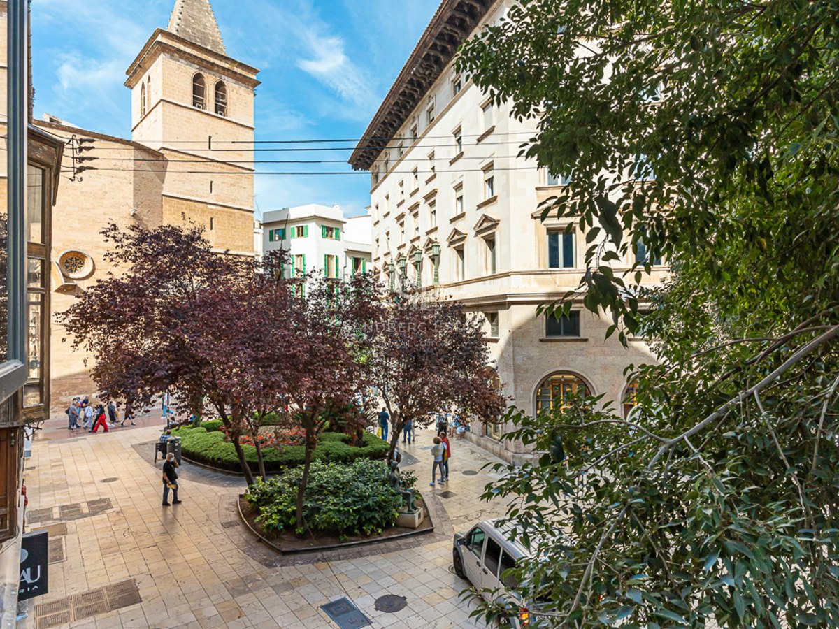 Pedestrian plaza with trees, stone buildings, and a church tower under a clear blue sky.