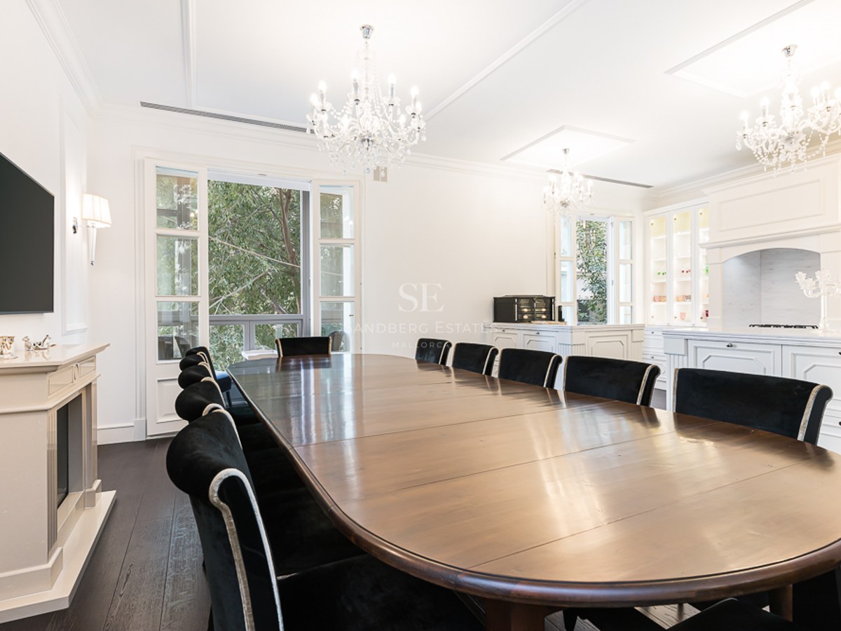 Large wooden dining table with black velvet chairs under crystal chandeliers in a bright, white room with a fireplace.