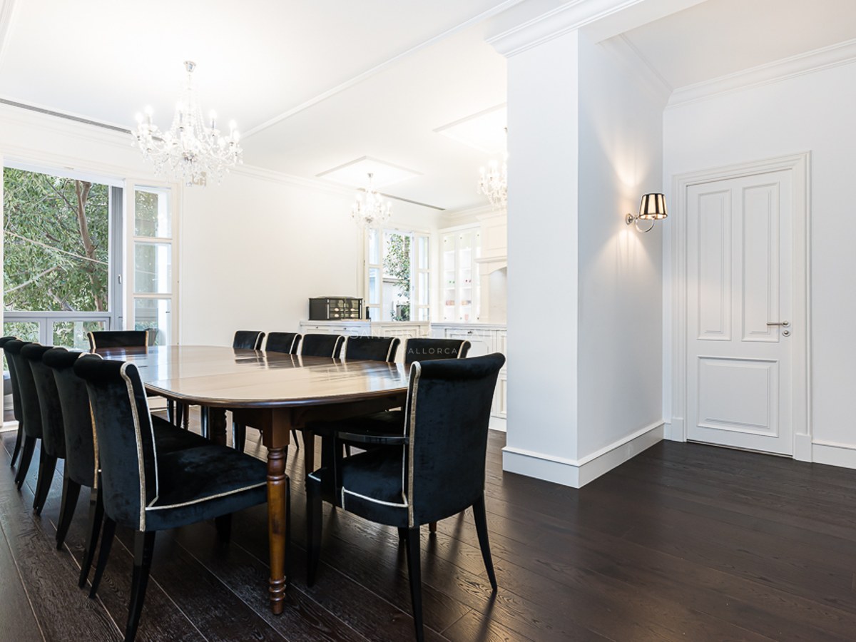 A large dark wood dining table with black velvet chairs under crystal chandeliers in a bright white room.