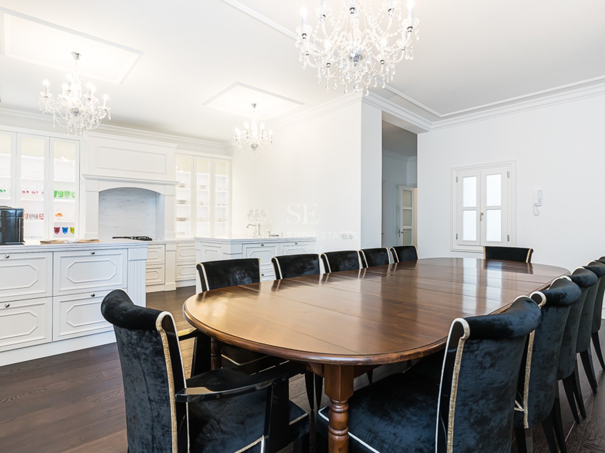 Large oval dark wood dining table surrounded by black velvet chairs under ornate crystal chandeliers in a white room.