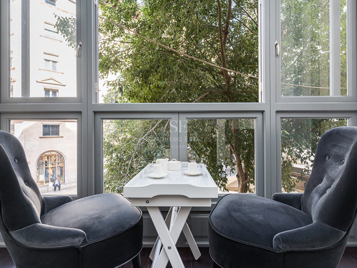 Two charcoal velvet armchairs facing a large window with a white tea table in between, overlooking city trees.