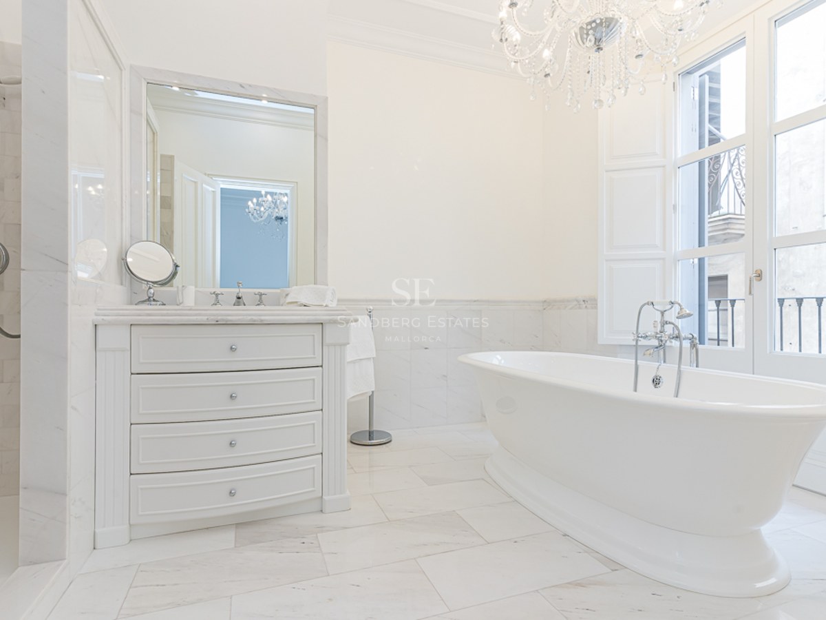 Luxury white marble bathroom featuring a freestanding tub, crystal chandelier, and large window with natural light.
