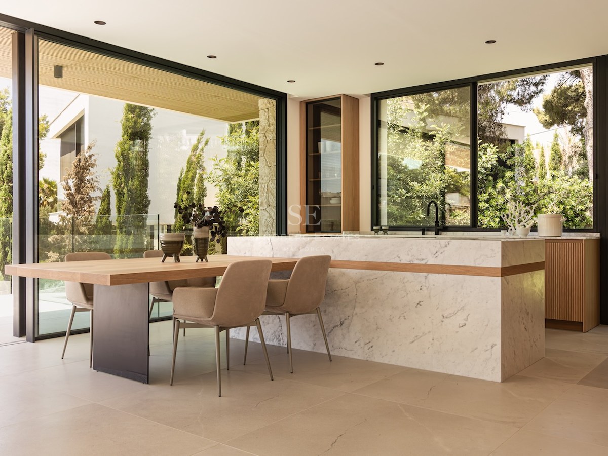 Salle à manger lumineuse avec table en bois, chaises beiges, îlot en marbre et grandes baies vitrées sur le jardin.