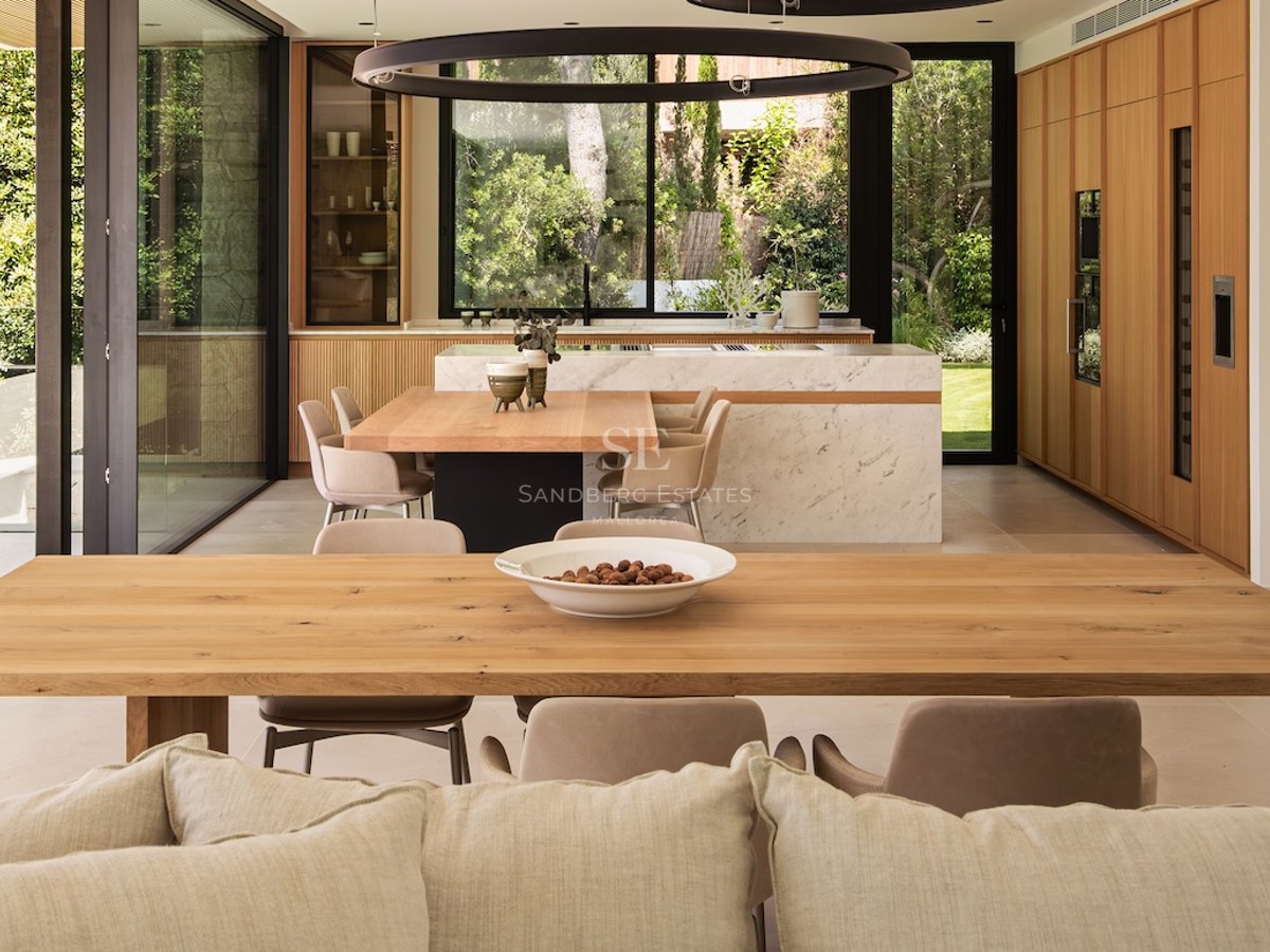 Salle à manger ouverte avec tables en bois, îlot en marbre et grandes vitres donnant sur un jardin verdoyant.