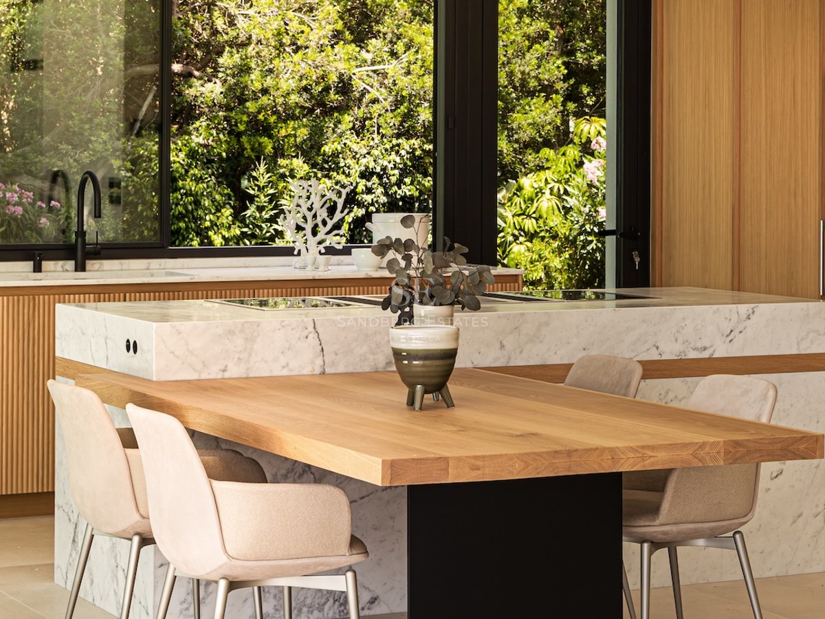 Modern kitchen featuring a white marble island with integrated wooden dining table and large windows showing lush gardens.