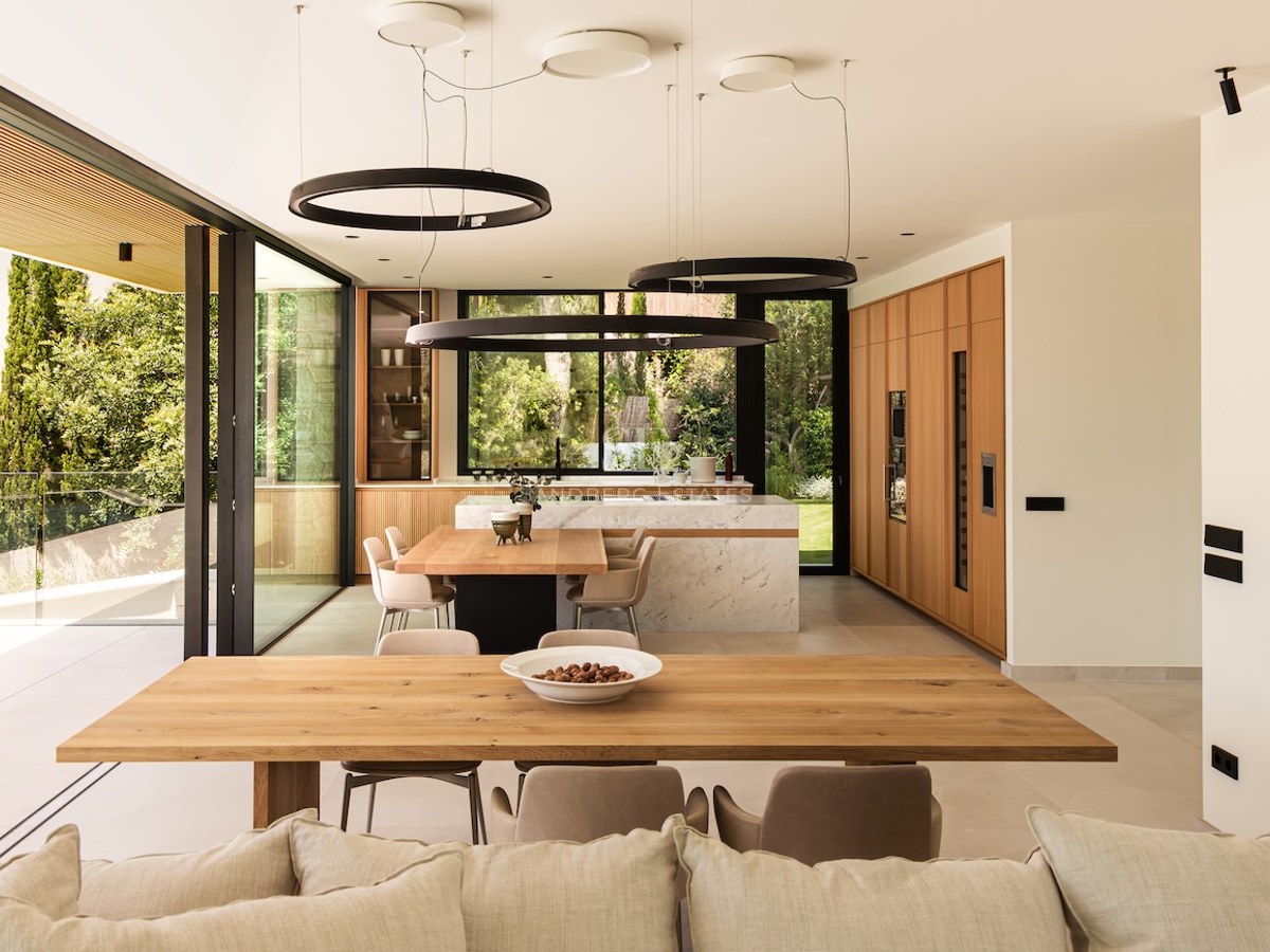 Salle à manger lumineuse avec table en bois, îlot en marbre, luminaires circulaires noirs et vue sur le jardin.