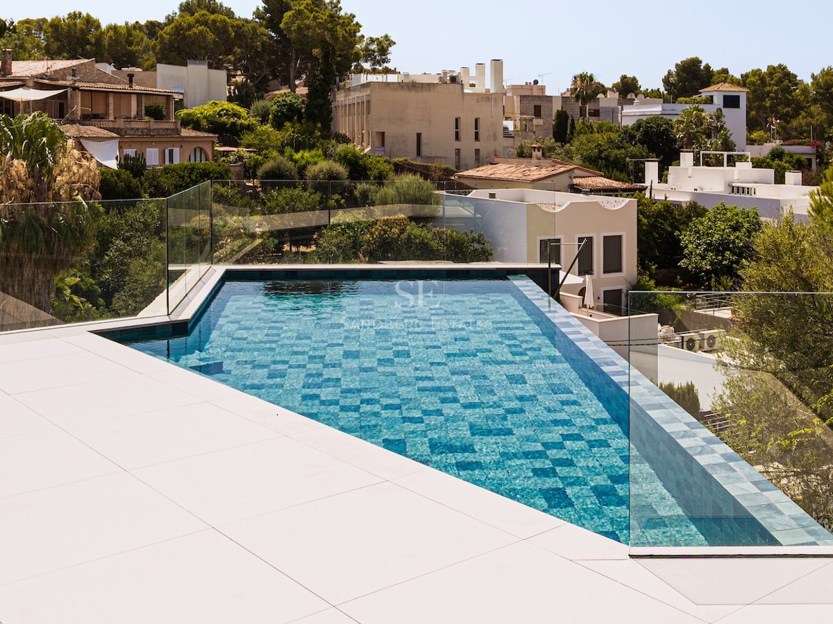 Piscine à débordement aux carreaux bleus avec garde-corps en verre surplombant une ville méditerranéenne verdoyante.