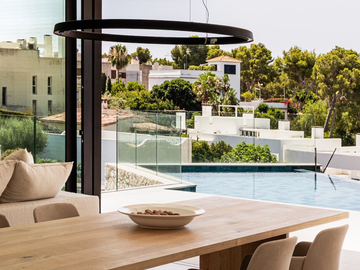 Salle à manger lumineuse avec table en bois et vue sur une piscine et un paysage verdoyant à travers de grandes vitres.
