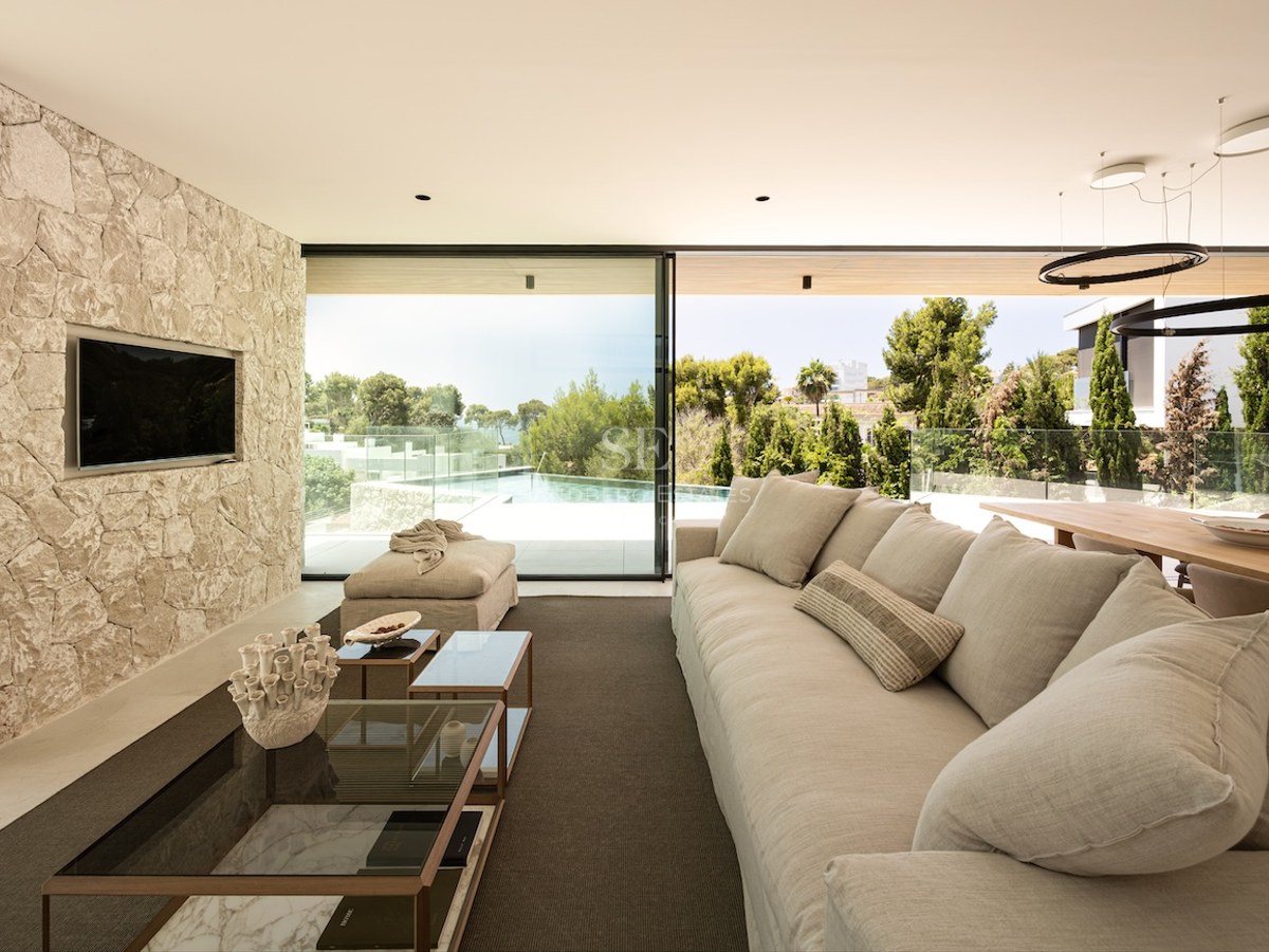 Salon moderne avec grand canapé beige, mur en pierre naturelle et baies vitrées ouvrant sur une piscine à débordement.