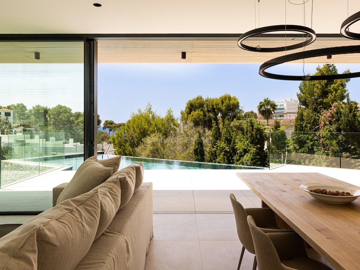 Salon lumineux avec canapé beige ouvrant sur une terrasse avec piscine à débordement et vue sur la nature.