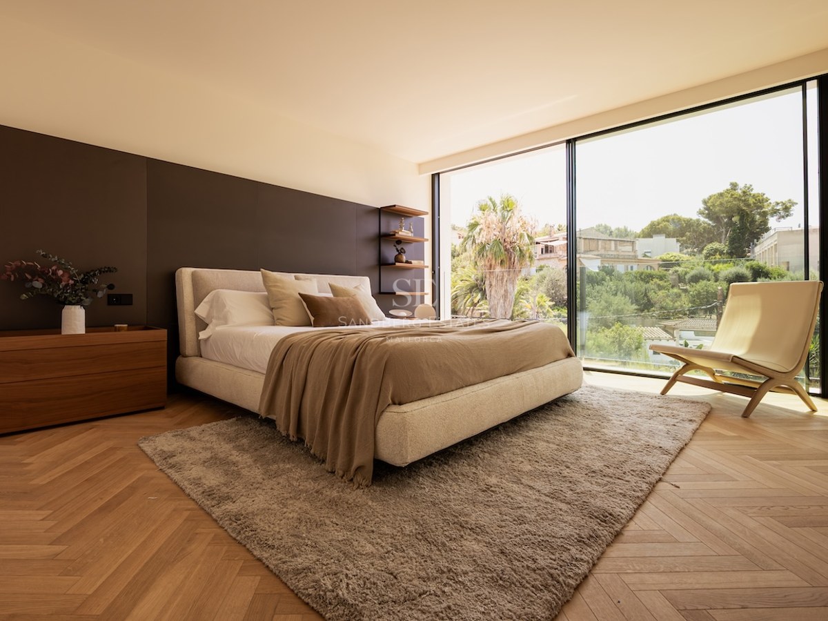 Chambre moderne avec parquet en chevrons, grand lit, fauteuil design et baie vitrée ouvrant sur un jardin tropical.