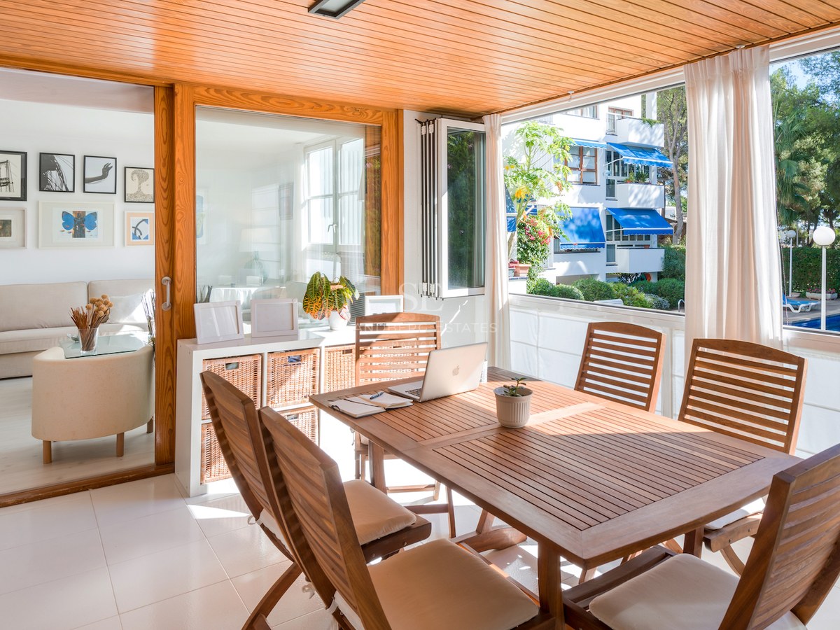 Terrasse fermée en bois avec table à manger et ordinateur, donnant sur une piscine et un jardin.