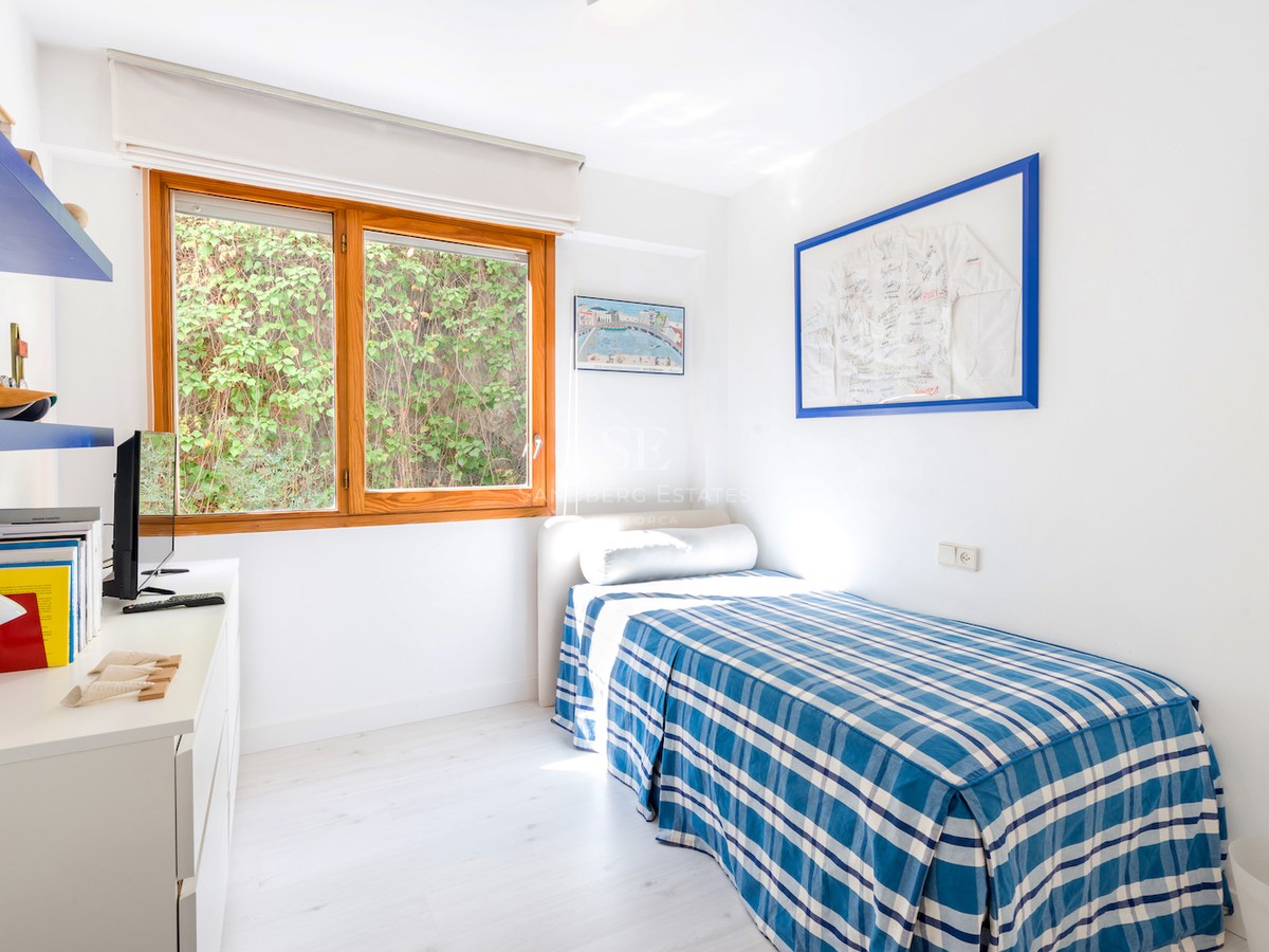 Chambre simple lumineuse avec un lit à carreaux bleus, un bureau blanc et une grande fenêtre en bois.