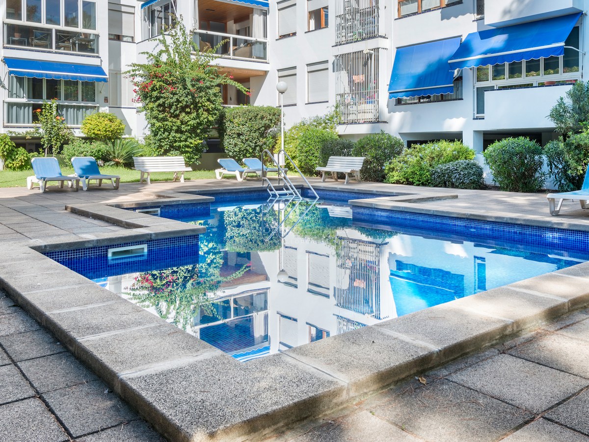 Piscine rectangulaire bleue entourée d'une terrasse en pierre avec des transats devant un immeuble blanc.