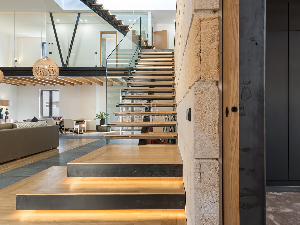 Modern open-plan living room featuring a wooden floating staircase with LED lights, glass railings, and a mezzanine.