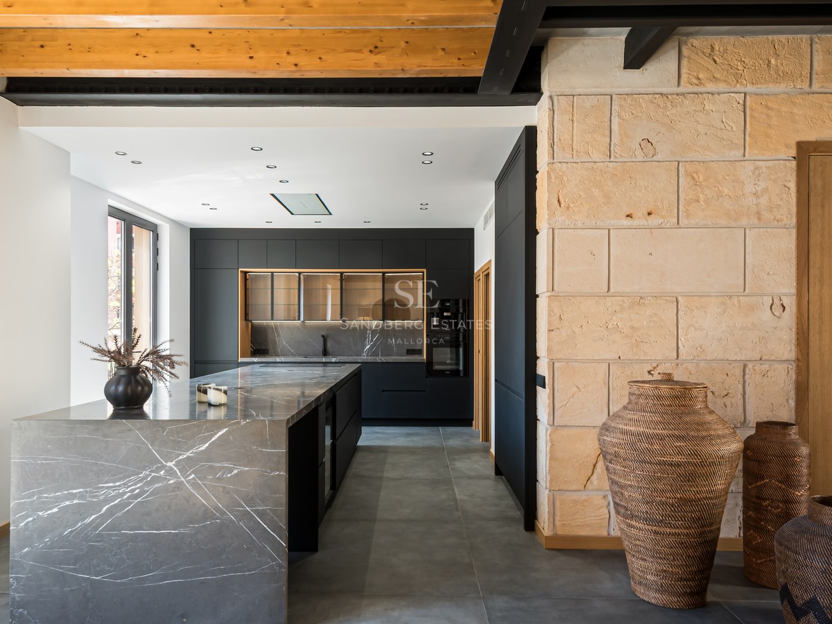 A contemporary kitchen featuring a grey marble island, black cabinetry, exposed wooden ceiling beams, and a stone wall.