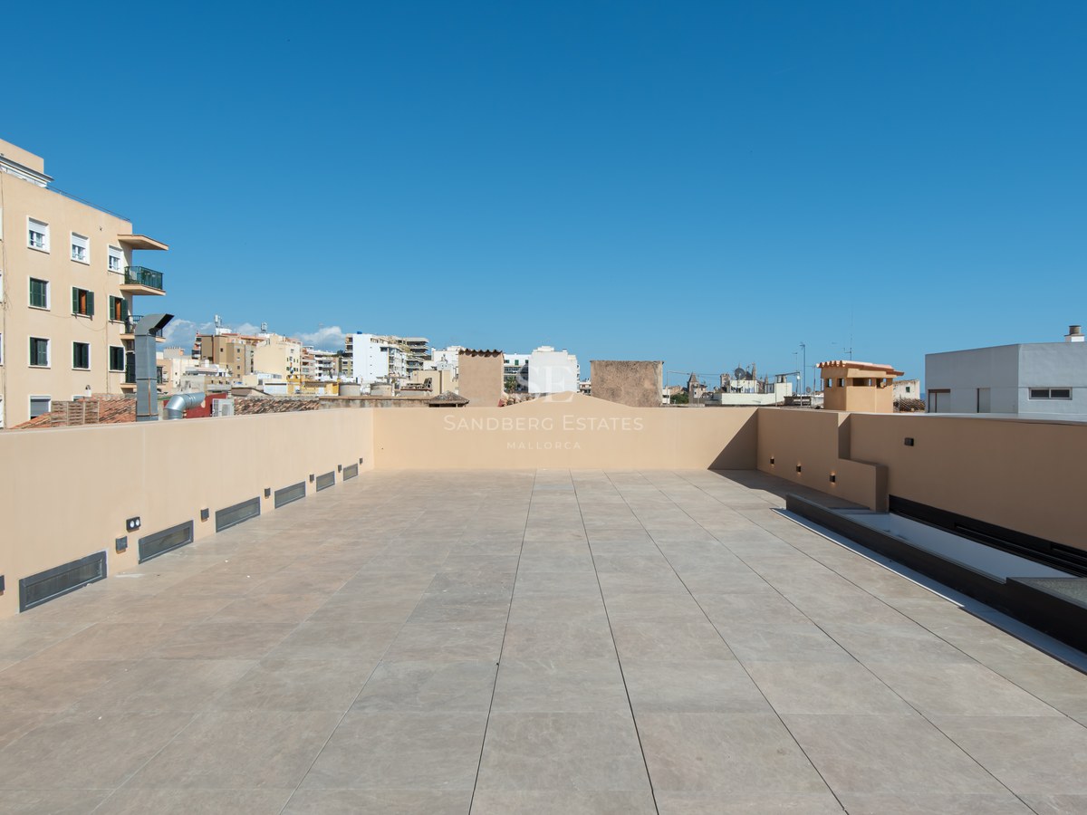 Spacious rooftop terrace with beige tiles and modern wall lighting under a clear blue sky.