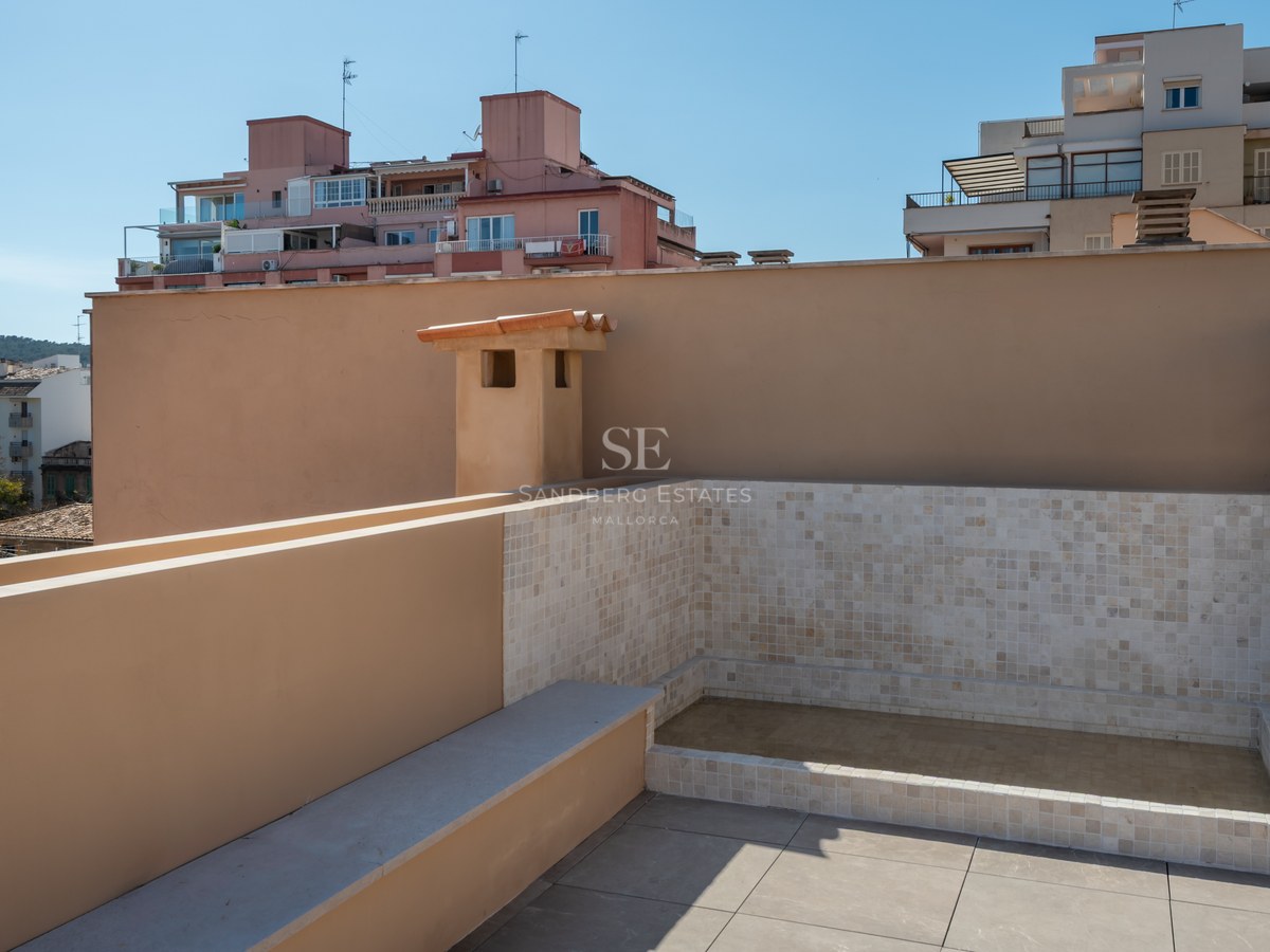 A modern rooftop terrace featuring a tiled plunge pool, beige stone flooring, and views of city buildings under a blue sky.