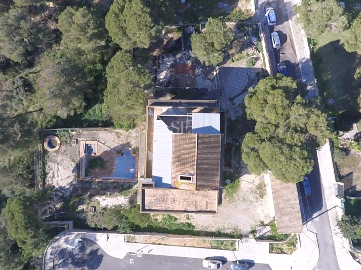 Top-down aerial view of a house with tiled roof, empty swimming pool, and surrounding pine trees.