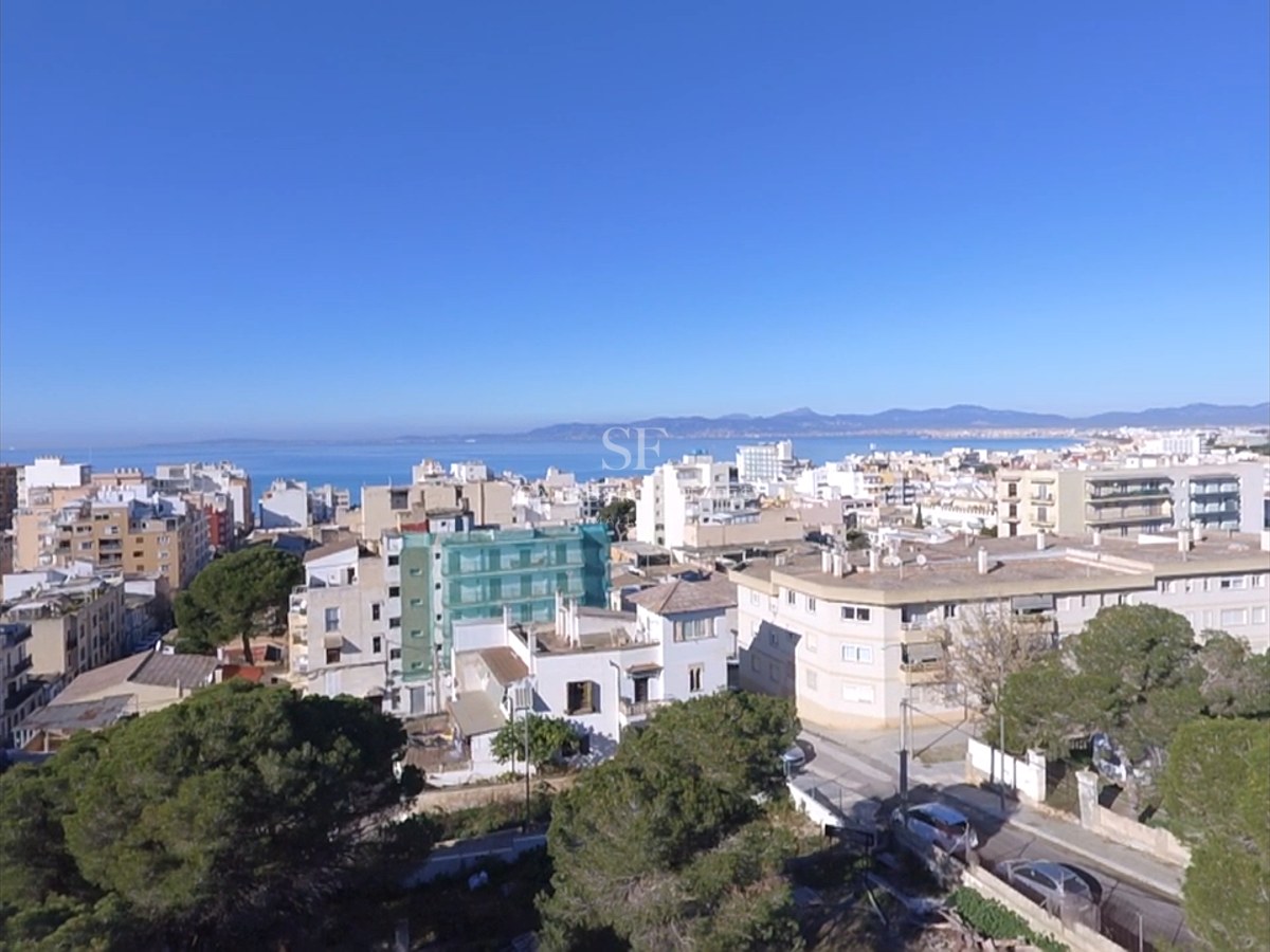 Elevated view of a Mediterranean coastal city with white buildings, green trees, and the blue sea on the horizon.