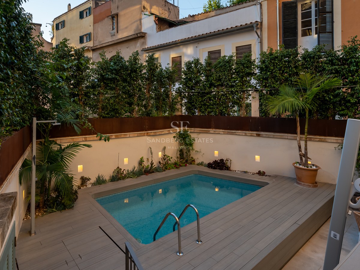 Modern rectangular pool with grey decking, surrounded by white walls with integrated lighting and lush greenery.