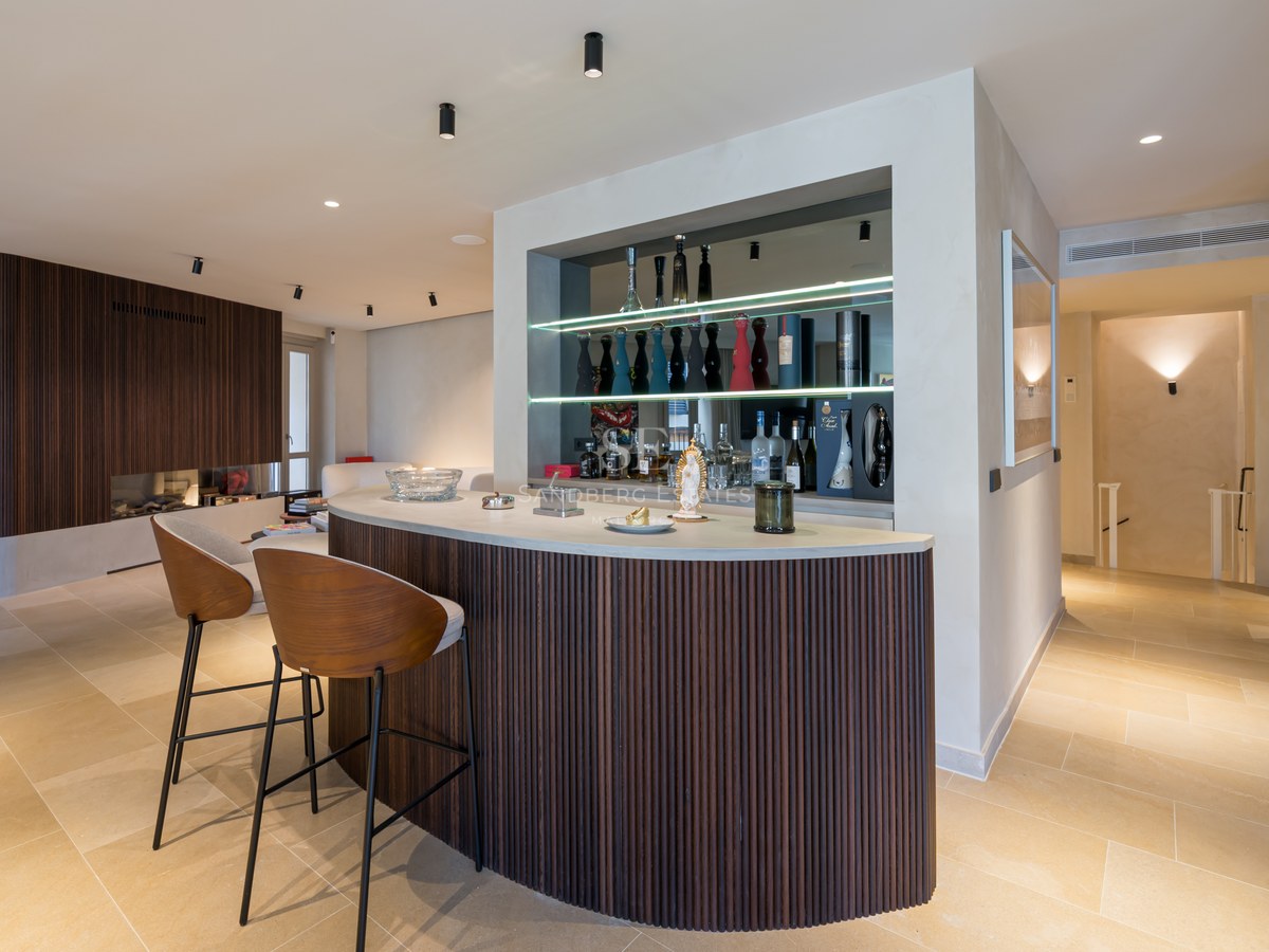 Curved home bar with dark wood slats, two designer stools, and backlit shelves with premium liquor bottles.