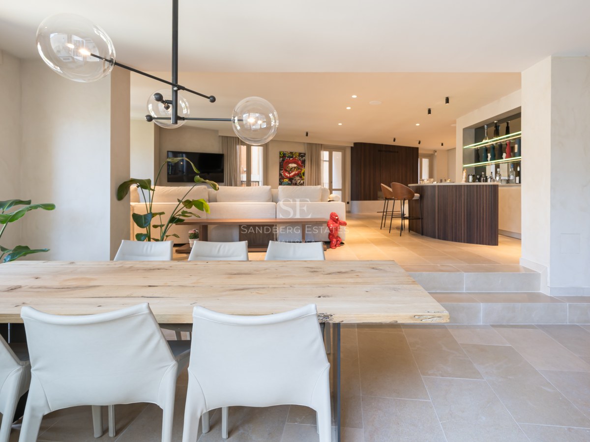 A spacious modern open-plan room featuring a wooden dining table, white chairs, glass globe pendants, and a bar area.