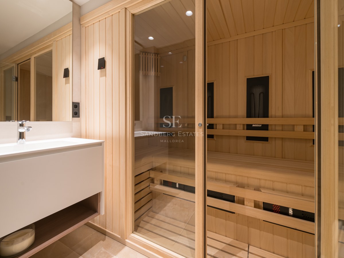 Light wood sauna with glass doors next to a sleek white bathroom vanity and mirror.