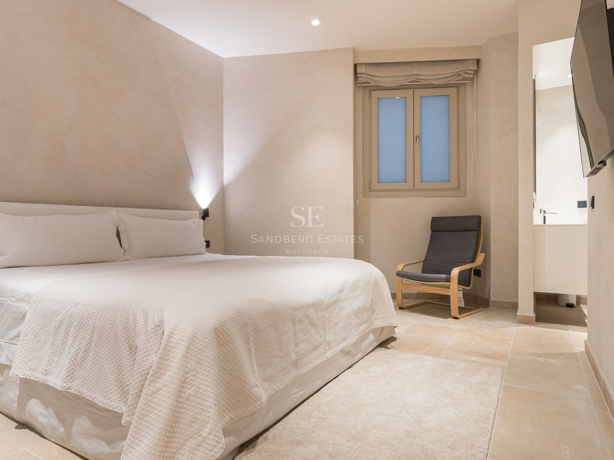A serene bedroom with a large white bed, a grey designer chair, stone flooring, and soft beige walls.