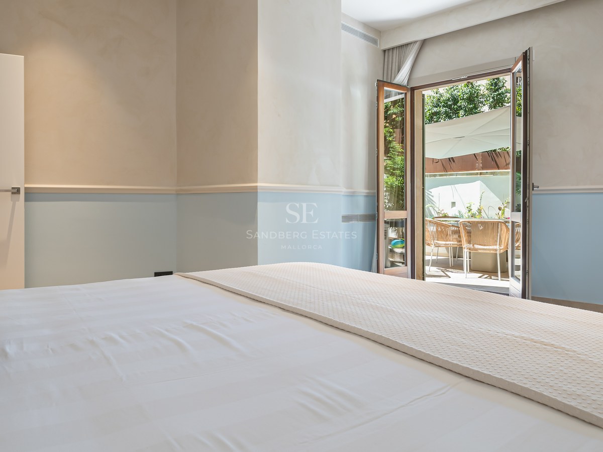 Bedroom with white linens and light blue walls looking out onto a sunny terrace through open French doors.