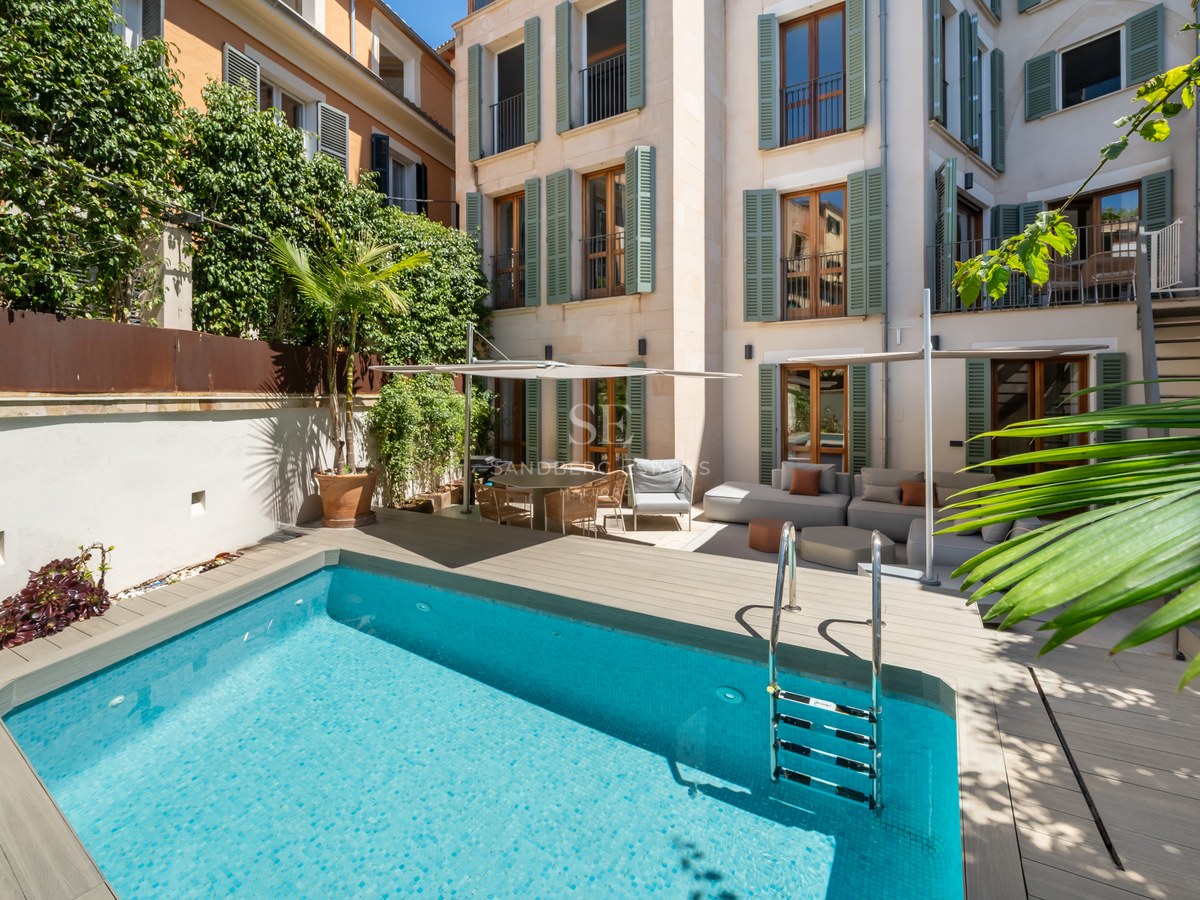 Light blue pool in a courtyard with wooden decking, outdoor sofas, and a building facade with green shutters.