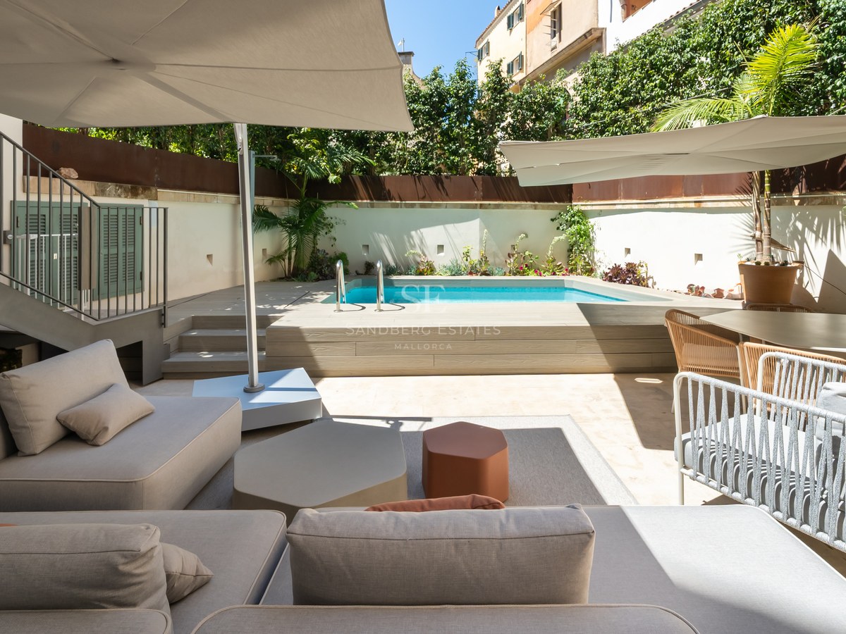 Modern terrace featuring a turquoise pool, wooden deck, grey sofas, and sun umbrellas under a clear sky.