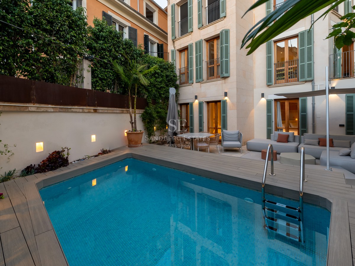 Modern private pool area with wooden decking, grey outdoor sofas, and a Mediterranean-style building with green shutters.