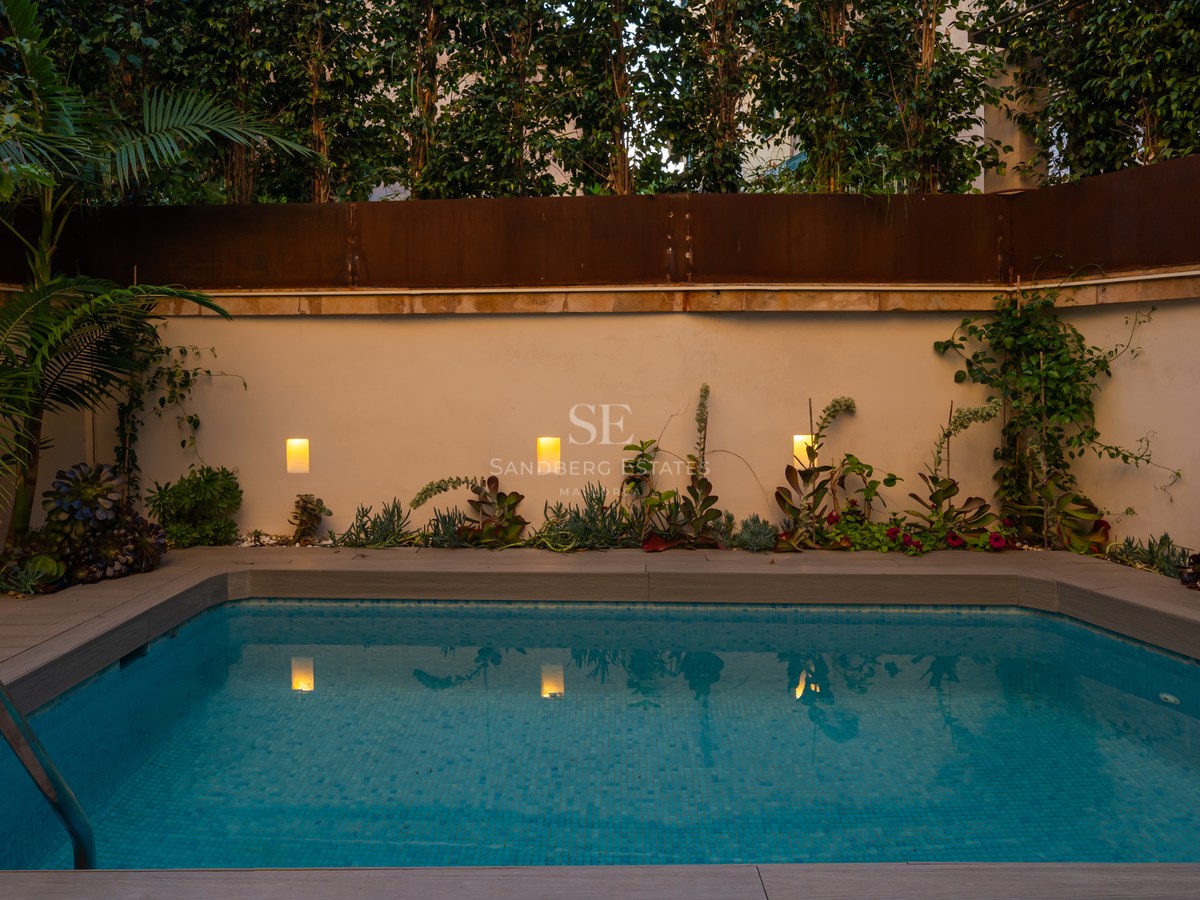Small rectangular blue-tiled pool at dusk with warm wall lights, wood-effect decking, and lush greenery.