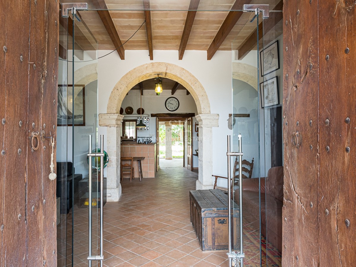 Large rustic wooden doors open to a grand foyer with a stone archway, terracotta floors, and wooden ceiling beams.