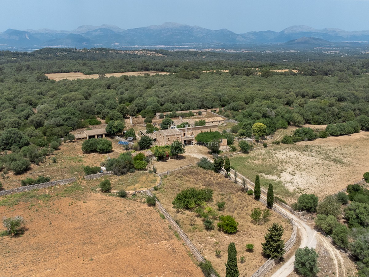 Aerial view of a large stone estate surrounded by forests and fields with mountains in the background under a clear sky.