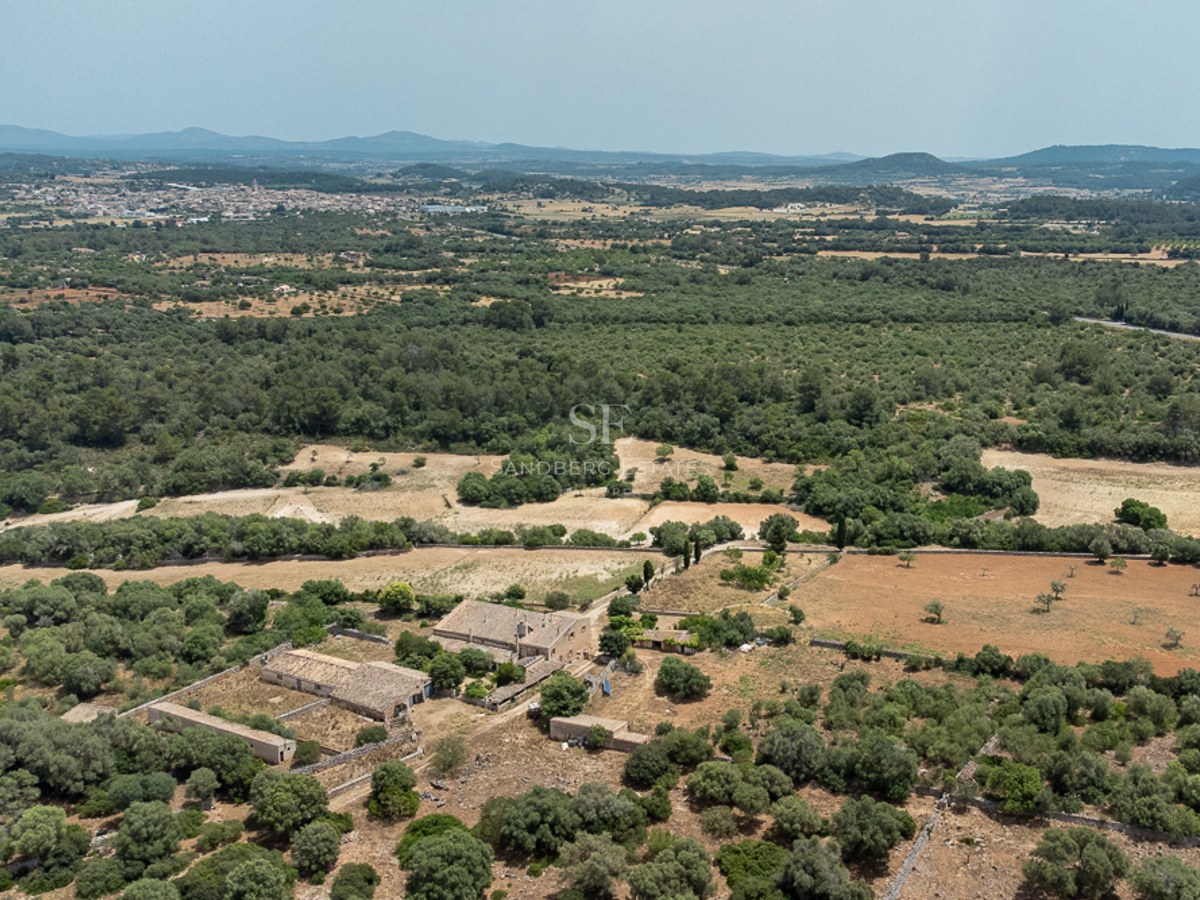 High-angle drone shot of a large stone farmhouse surrounded by lush greenery and vast fields under a clear sky.