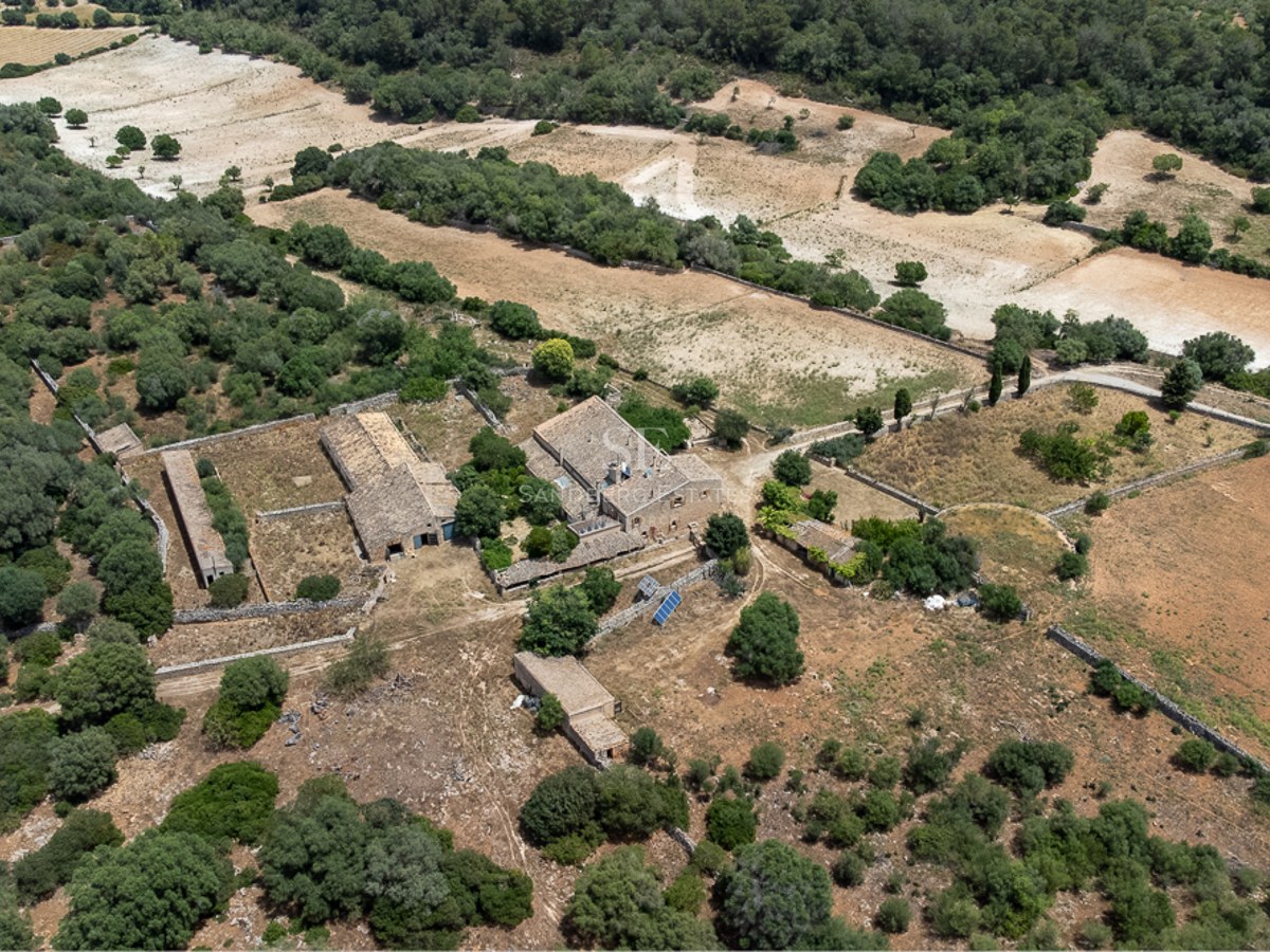 Aerial drone shot of a rustic stone farmhouse complex with multiple outbuildings surrounded by dry fields and green trees.