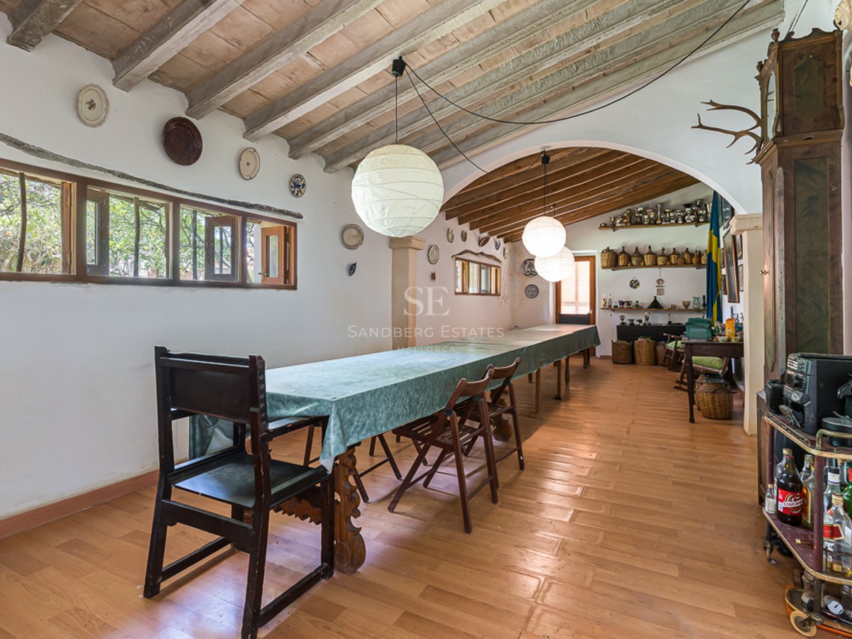 A long dining table with dark wood chairs in a rustic room with exposed beam ceilings and white plaster walls.
