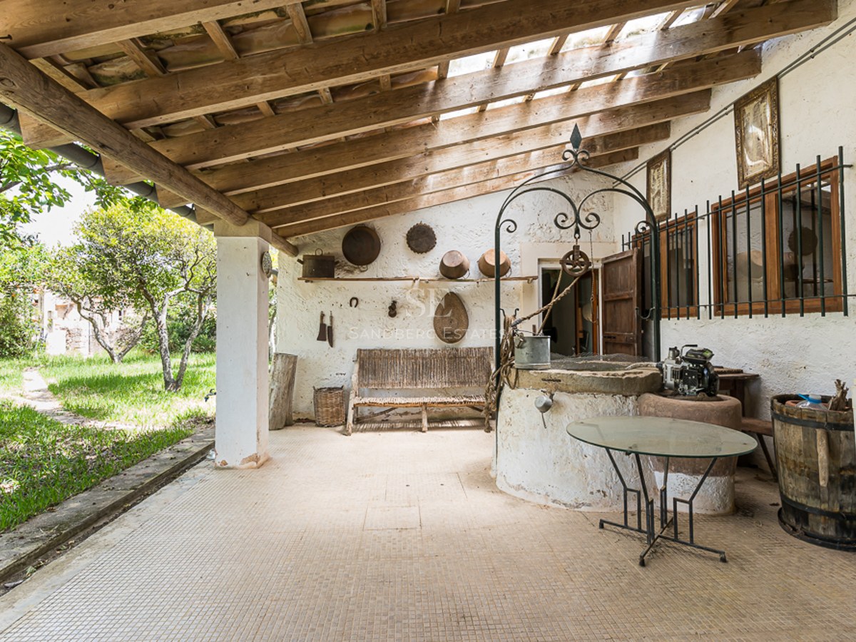 Covered terrace featuring an antique stone well, exposed wooden beams, and rustic decor overlooking a green garden.