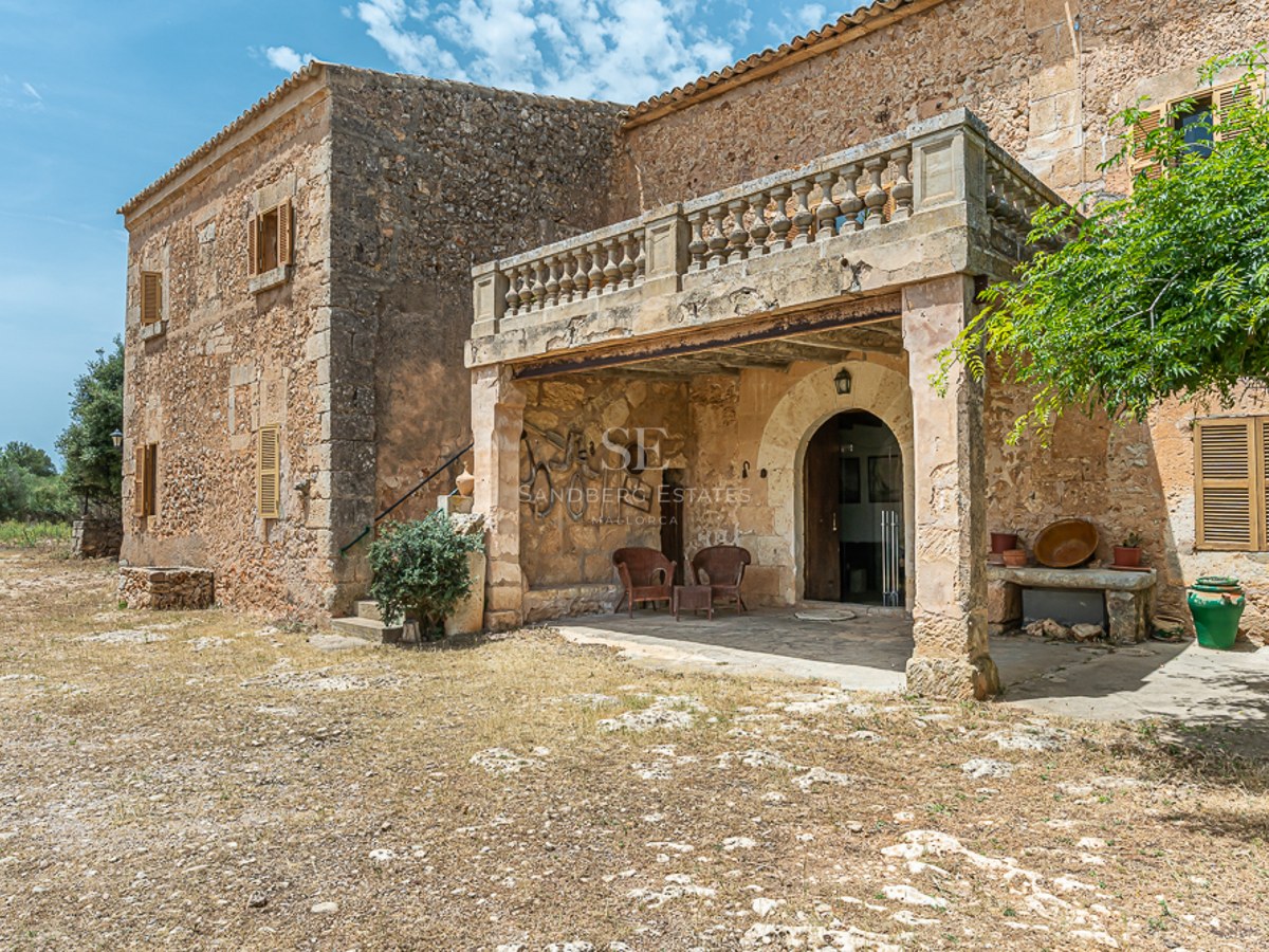 Rustic stone building with an arched entrance, stone pillars supporting a balustrade balcony under a blue sky.