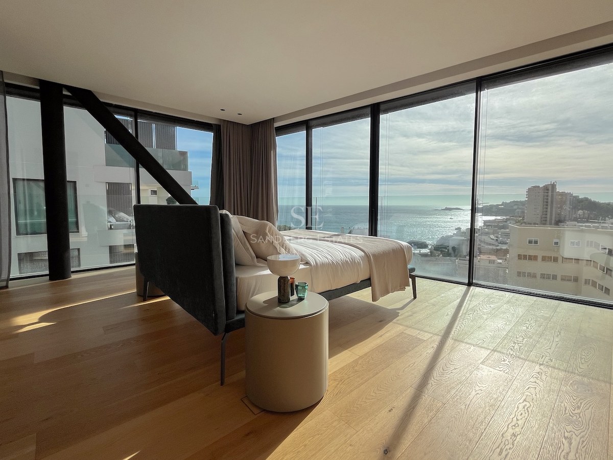 Modernes Schlafzimmer mit bodentiefen Glaswänden, hellem Eichenparkett und Blick auf das Meer und die Stadtküste.