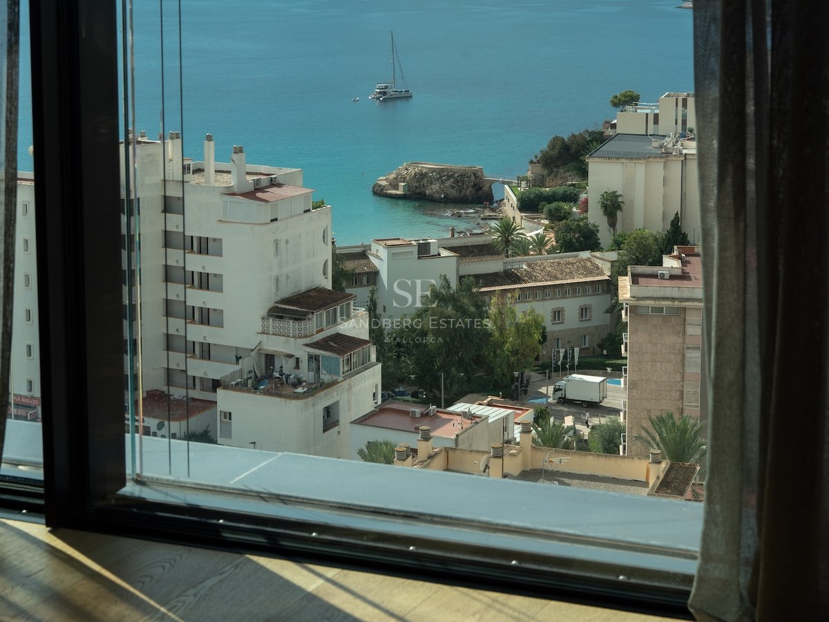 View from a modern window overlooking a turquoise bay with a sailboat and white Mediterranean buildings.