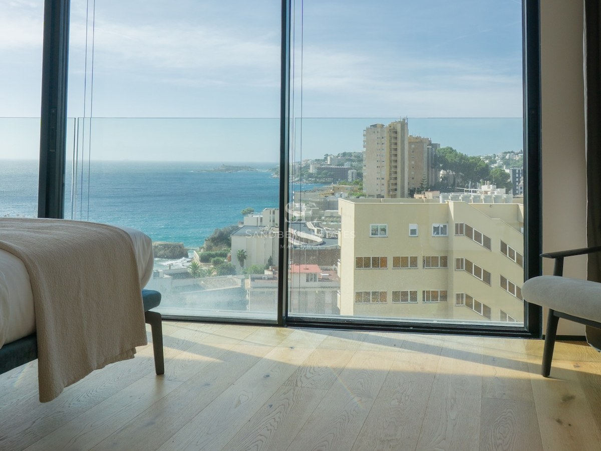 Interior of a minimalist bedroom featuring floor-to-ceiling windows overlooking the Mediterranean coast and city.