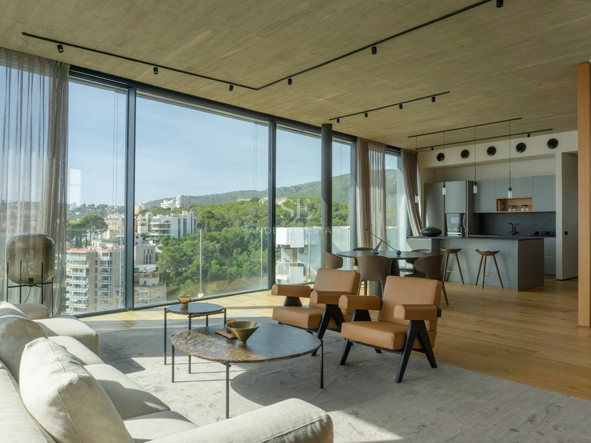 Modern open-plan living room with floor-to-ceiling windows overlooking a Mediterranean city and green hills.